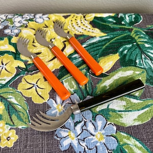 May include: A set of vintage serving forks with stainless steel tines and orange handles, along with a larger fork with a black handle. The forks are arranged on a floral patterned fabric.
