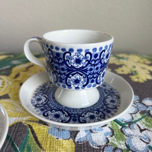 May include: A white teacup and saucer set with a blue floral pattern. The teacup has a handle and sits on a pedestal base. The saucer mirrors the cup's design. The set is on a floral patterned tablecloth.