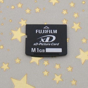 Puede incluir: Una tarjeta xD-Picture Card negra de Fujifilm con texto blanco. La tarjeta tiene el logotipo de Fujifilm, el logotipo xD y el texto "xD-Picture Card" y "M 1GB". La tarjeta es rectangular con un lado con muescas.