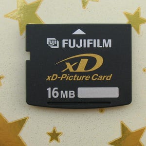 Puede incluir: Una tarjeta xD-Picture Fujifilm negra con letras doradas y el texto "16 MB". La tarjeta tiene forma rectangular con un borde dentado. El fondo es claro con formas de estrellas doradas.