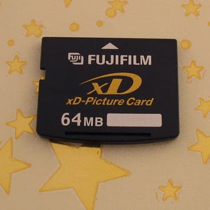 Puede incluir: Una tarjeta Fujifilm xD-Picture negra con texto blanco y un logotipo dorado. La tarjeta dice "xD-Picture Card" y "64 MB". El fondo es de color amarillo claro con formas de estrellas doradas.