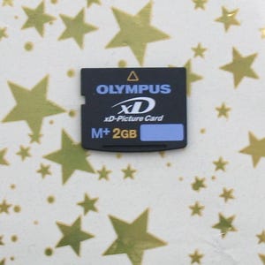 Puede incluir: Una tarjeta xD-Picture Olympus negra con el texto "OLYMPUS", "xD", "xD-Picture Card" y "M+ 2GB". La tarjeta tiene una franja azul y está sobre un fondo con formas de estrellas doradas.