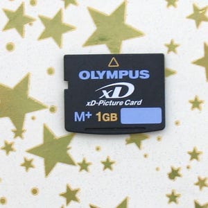 Puede incluir: Una tarjeta Olympus xD-Picture negra con el texto "OLYMPUS", "xD-Picture Card" y "M+ 1GB". La tarjeta está sobre un fondo blanco con formas de estrellas doradas.