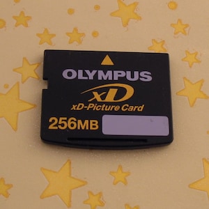 Puede incluir: Tarjeta xD-Picture Olympus negra con el texto "OLYMPUS", "xD" y "256MB". La tarjeta tiene forma rectangular con un borde dentado. El fondo es de color amarillo claro con formas de estrellas doradas.