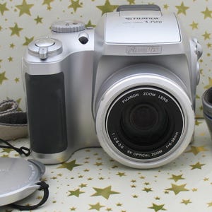 May include: Silver Fujifilm FinePix S 2500HD digital camera with a lens cap, lens hood, and camera strap. The camera has a black grip and a zoom lens. The background is a white and gold star pattern.