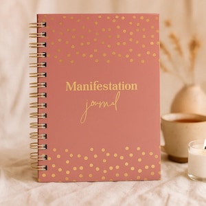 May include: A pink spiral-bound journal with gold polka dots and gold lettering that reads "Manifestation journal." The journal is open, revealing blank pages. A candle and a cup are in the background.