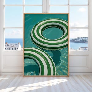 May include: A framed print featuring two green and white striped inflatable pool floats floating in turquoise water. The artwork is displayed in a room with a large window overlooking a coastal scene.