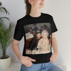 Puede incluir: Camiseta negra con una imagen vintage de tres personas en una mesa. La imagen de la camiseta incluye a un hombre con un sombrero de Kings. La camiseta la lleva una persona en un entorno bien iluminado.