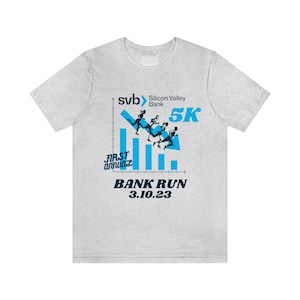 May include: Light gray t-shirt featuring a blue graphic of runners on a downward-sloping graph. The text includes "svb Silicon Valley Bank 5K", "First Annual", and "BANK RUN 3.10.23". The shirt is a crew neck style.