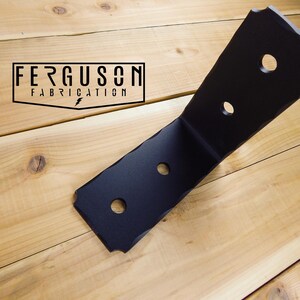 May include: A matte black L-shaped metal bracket with four circular mounting holes and textured, uneven edges. The bracket features the text FERGUSON FABRICATION printed in a black rectangular frame on the surface next to the metal object.