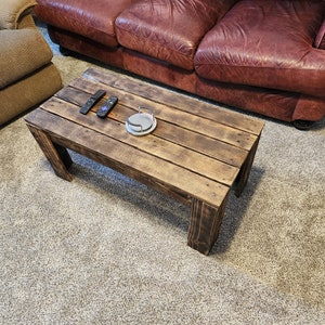 May include: A rustic wooden coffee table with a distressed brown finish. The table has a rectangular top with visible wood grain and is supported by four sturdy legs. Two remote controls and a round silver object are placed on the table.