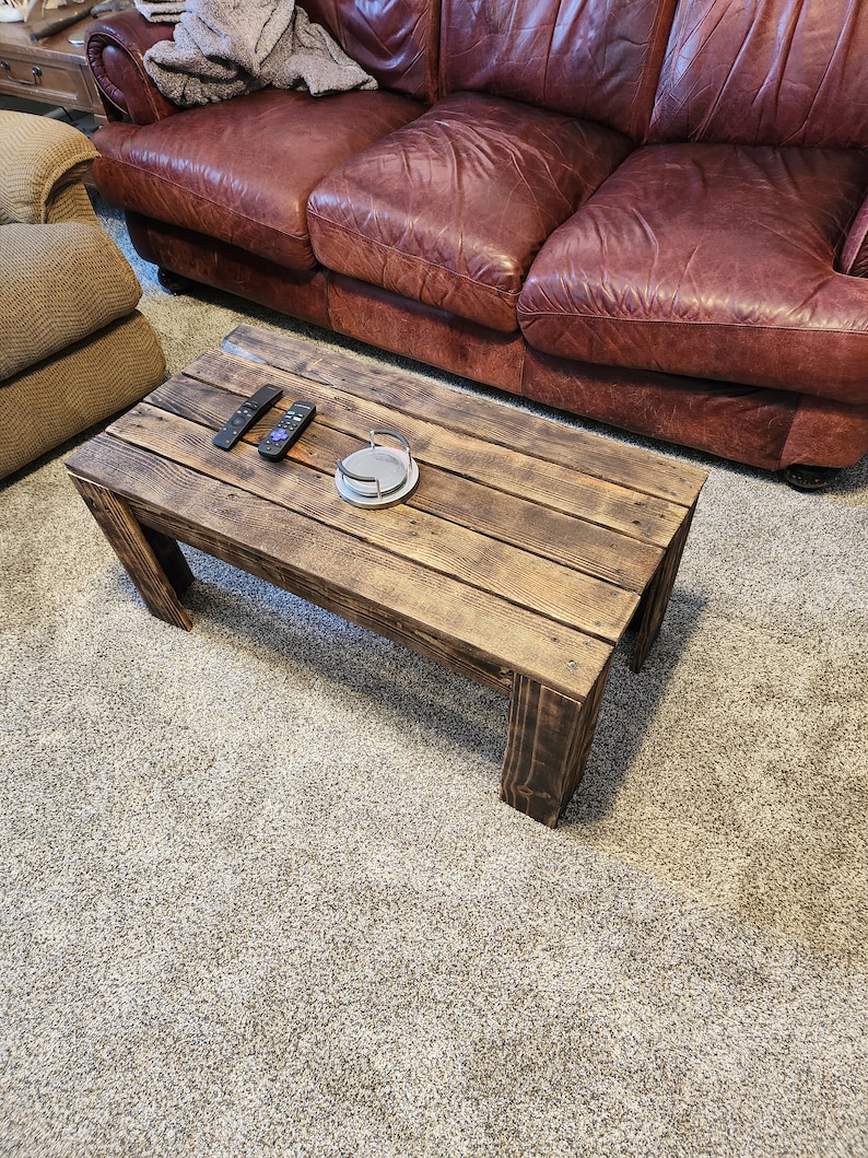 May include: A rustic wooden coffee table with a distressed brown finish. The table has a rectangular top with visible wood grain and is supported by four sturdy legs. Two remote controls and a round silver object are placed on the table.