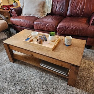 May include: A wooden coffee table with a lower shelf, a tray with a bowl, a remote control, and a small potted plant. The table is in front of a brown leather sofa with pillows.