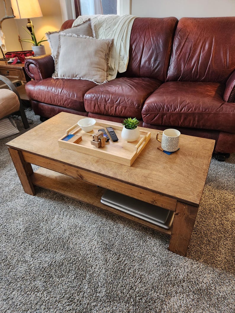 May include: A wooden coffee table with a lower shelf, a tray with a bowl, a remote control, and a small potted plant. The table is in front of a brown leather sofa with pillows.