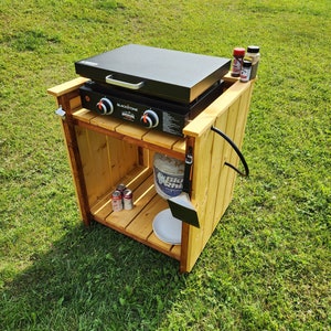 Puede incluir: Una estaci&oacute;n de cocina exterior de madera con una plancha Blackstone Adventure negra. La estaci&oacute;n tiene un tanque de propano, una esp&aacute;tula y varios recipientes de especias. La plancha tiene dos perillas de control y una tapa.