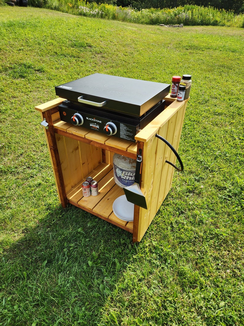 Puede incluir: Una estaci&oacute;n de cocina exterior de madera con una plancha Blackstone Adventure negra. La estaci&oacute;n tiene un tanque de propano, una esp&aacute;tula y varios recipientes de especias. La plancha tiene dos perillas de control y una tapa.