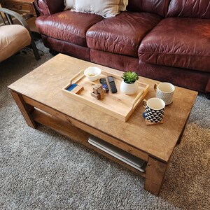 May include: A wooden coffee table with a light brown finish and a lower shelf. The table has a tray with a bowl, remote controls, and a small potted plant. Two mugs are on the table, one with a black and white geometric pattern and the other with a black and white striped pattern.