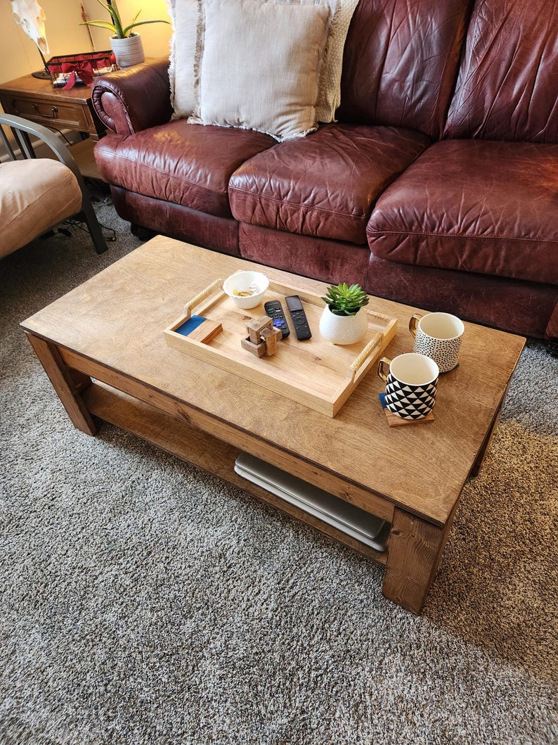 May include: A wooden coffee table with a light brown finish and a lower shelf. The table has a tray with a bowl, remote controls, and a small potted plant. Two mugs are on the table, one with a black and white geometric pattern and the other with a black and white striped pattern.
