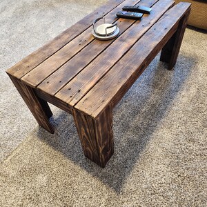 May include: A rustic wooden coffee table with a dark brown stain. The table has a rectangular top and four legs. The table is made of reclaimed wood and has a distressed look.