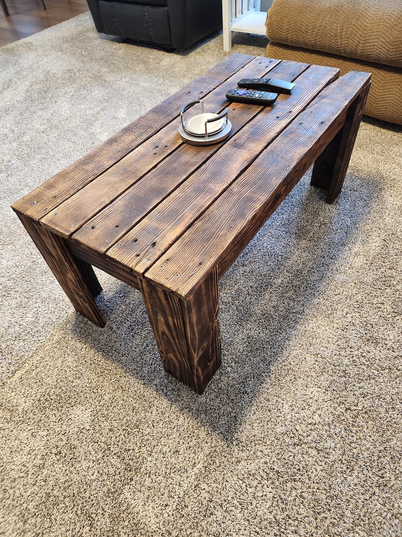 May include: A rustic wooden coffee table with a dark brown stain. The table has a rectangular top and four legs. The table is made of reclaimed wood and has a distressed look.