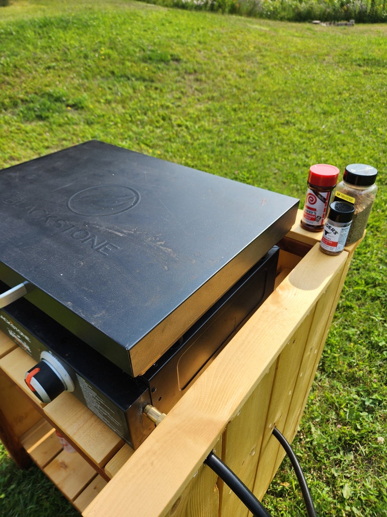 Puede incluir: Una plancha Blackstone negra sobre un soporte de madera, colocada sobre c&eacute;sped. Tres botes de especias est&aacute;n en el estante de madera. La plancha tiene un logotipo circular y la palabra "BLACKSTONE".