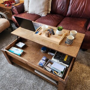 May include: A wooden lift-top coffee table with a lower shelf. The table is open, revealing a laptop, a tray with a small bowl, a remote control, a small plant, and a wooden puzzle. The lower shelf is filled with various items, including a book, a box of batteries, and a roll of tape.