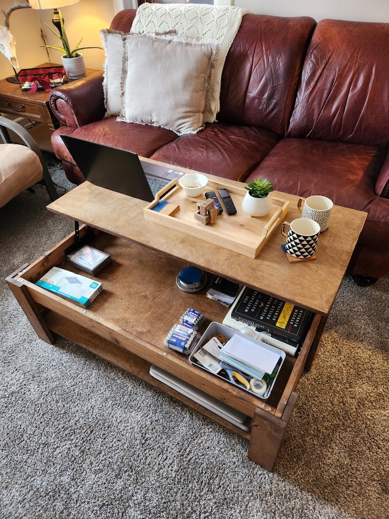 May include: A wooden lift-top coffee table with a lower shelf. The table is open, revealing a laptop, a tray with a small bowl, a remote control, a small plant, and a wooden puzzle. The lower shelf is filled with various items, including a book, a box of batteries, and a roll of tape.