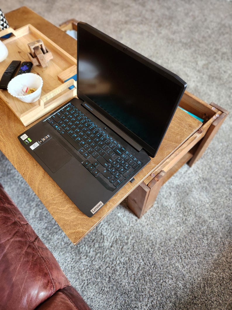 May include: A black laptop computer with a blue backlit keyboard is open on a wooden table. The laptop is on a wooden table with a tray on the left side of the table.