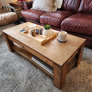 May include: A wooden coffee table with a lower shelf. The table has a natural wood finish and is sitting on a gray carpet. There is a tray with a small plant, a mug, and a few remote controls on the table.