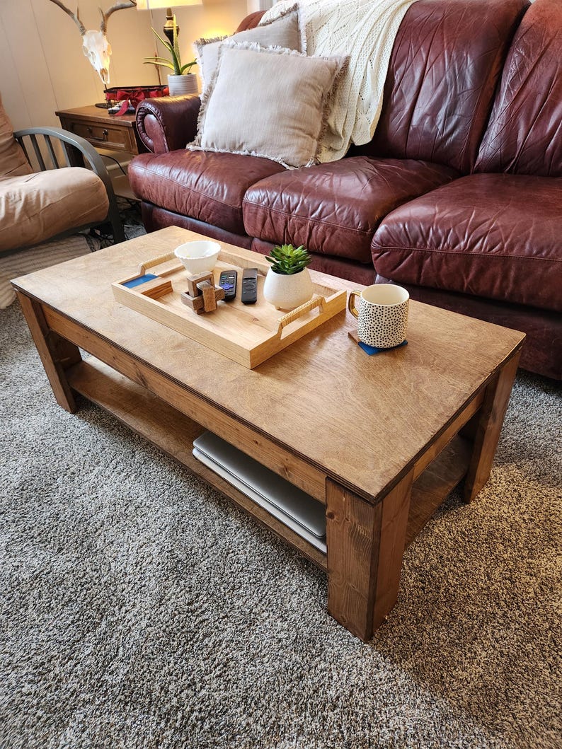 May include: A wooden coffee table with a lower shelf. The table has a natural wood finish and is sitting on a gray carpet. There is a tray with a small plant, a mug, and a few remote controls on the table.