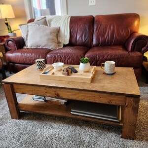 May include: A wooden coffee table with a lower shelf. The table has a natural wood finish and is sitting on a gray carpet. A leather sofa is in the background.