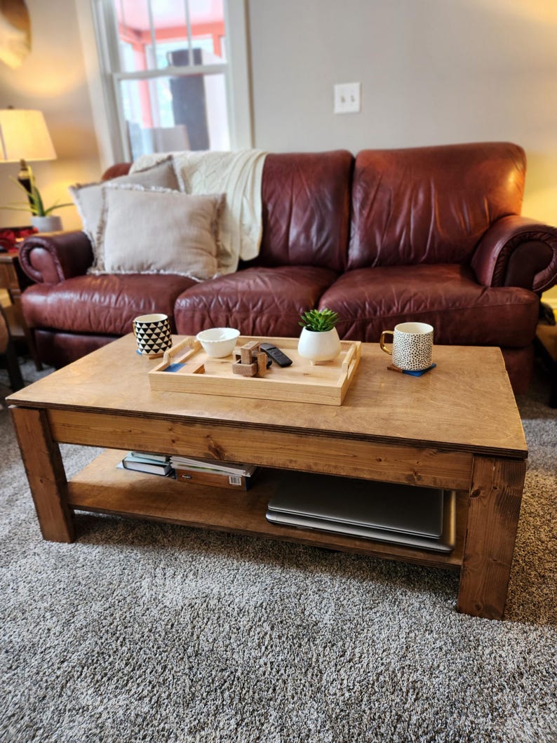 May include: A wooden coffee table with a lower shelf. The table has a natural wood finish and is sitting on a gray carpet. A leather sofa is in the background.