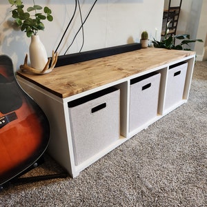 Peut inclure: Étagère de rangement en bois blanc avec trois bacs de rangement en tissu. L'étagère a un dessus en bois et est posée sur un tapis.