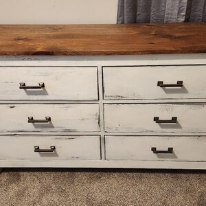 May include: White dresser with six drawers and a wood top. The drawers have black metal handles.