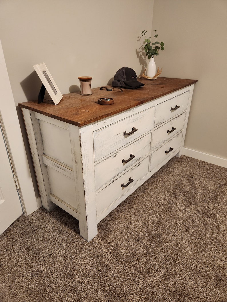 May include: White dresser with six drawers and black hardware. The dresser has a distressed white finish and a wood top. The dresser is sitting on a beige carpet.