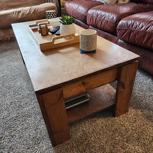 May include: A wooden coffee table with a lower shelf. The table has a natural wood finish and is sitting on a beige carpet. There are a few items on the table, including a tray, a plant, and two mugs.
