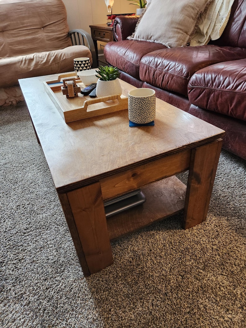May include: A wooden coffee table with a lower shelf. The table has a natural wood finish and is sitting on a beige carpet. There are a few items on the table, including a tray, a plant, and two mugs.