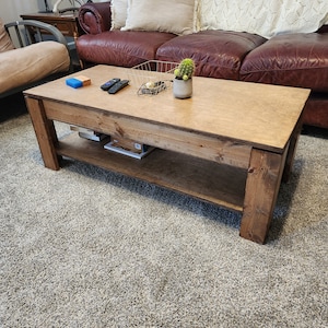 May include: A wooden coffee table with a natural finish and a lower shelf. The table has a rectangular top and is sitting on a light gray carpet. There are a few items on the table, including a small potted plant, a wire basket, and a couple of remote controls.