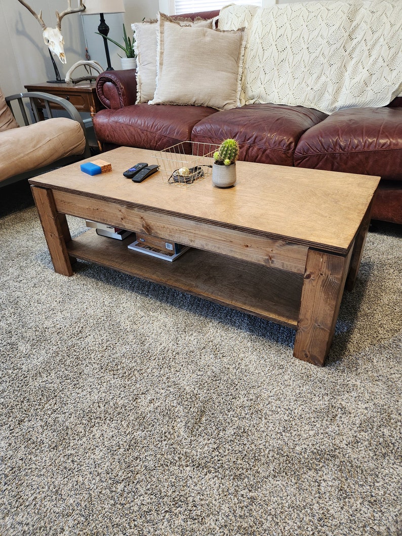 May include: A wooden coffee table with a natural finish and a lower shelf. The table has a rectangular top and is sitting on a light gray carpet. There are a few items on the table, including a small potted plant, a wire basket, and a couple of remote controls.