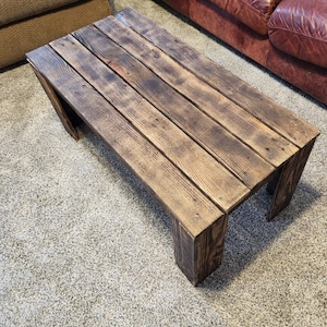 May include: A rustic wooden coffee table with a dark brown stain. The table has a rectangular top and four legs. The table is made of reclaimed wood and has a distressed look.