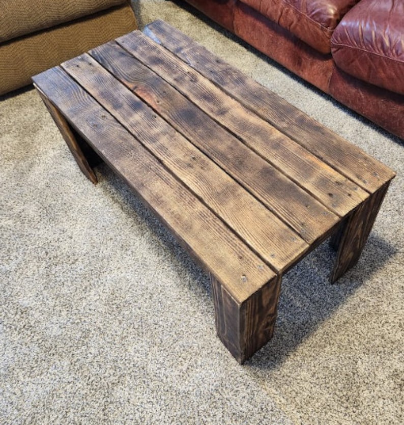 May include: A rustic wooden coffee table with a dark brown stain. The table has a rectangular top and four legs. The table is made of reclaimed wood and has a distressed look.