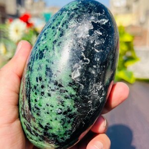 以下が含まれることがあります： 緑と黒が混ざった、磨かれた楕円形の石。石には斑点模様があり、光沢のある仕上がりです。ぼやけた背景には花と青い空があり、手で持たれています。