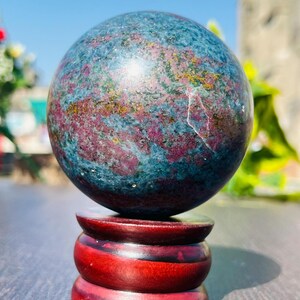 May include: A polished gemstone sphere with a mix of blue, red, and gold hues, resting on a tiered, dark red wooden stand. The sphere's surface has a marbled appearance. The stand is approximately 3 inches tall.