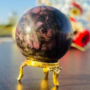 May include: A dark grey and pink sphere with a swirling pattern sits on a gold metal stand with three legs. The sphere is a polished stone.