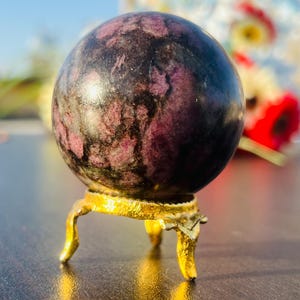 Peut inclure: Une sphère de couleur pourpre foncé et noire avec un motif tourbillonnant repose sur un support en métal doré à trois pieds. La sphère est sur une surface brun foncé.