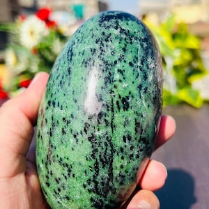 Peut inclure: Une pierre polie de forme ovale, avec une couleur de base verte vibrante et parsemée d'inclusions noires. La pierre est tenue dans une main, mettant en valeur sa surface lisse et tactile. L'arrière-plan est flou, avec des touches de verdure et de soleil.