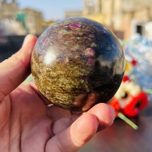 Könnte beinhalten: Eine polierte grüne und braune Kugel mit eingebetteten rosa Kristallen. Die Kugel ist glatt und hat eine glänzende Oberfläche.