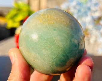 May include: A polished, spherical stone with a smooth surface, featuring a variegated color palette of soft seafoam green, muted teal, and subtle golden-brown inclusions, with a thin, light-colored natural fissure running across the upper portion.