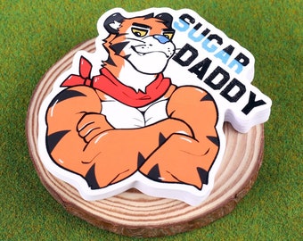 May include: A cartoon tiger with orange and black fur, wearing a red bandana, and the text "SUGAR DADDY" on a white background. The sticker is on a wooden slice, set against a green background.