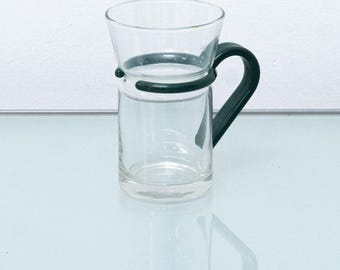 Op de afbeelding: Een heldere glazen beker met een zwarte kunststof handvat. Het handvat is bevestigd aan de beker met een zwarte kunststof ring.